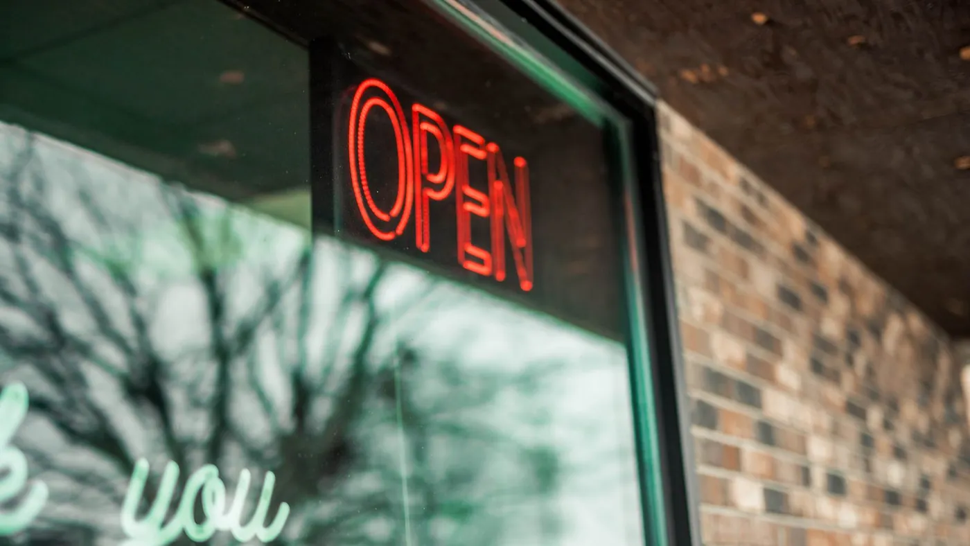 A glowing red neon Open sign in the window of a brick storefront at dusk.