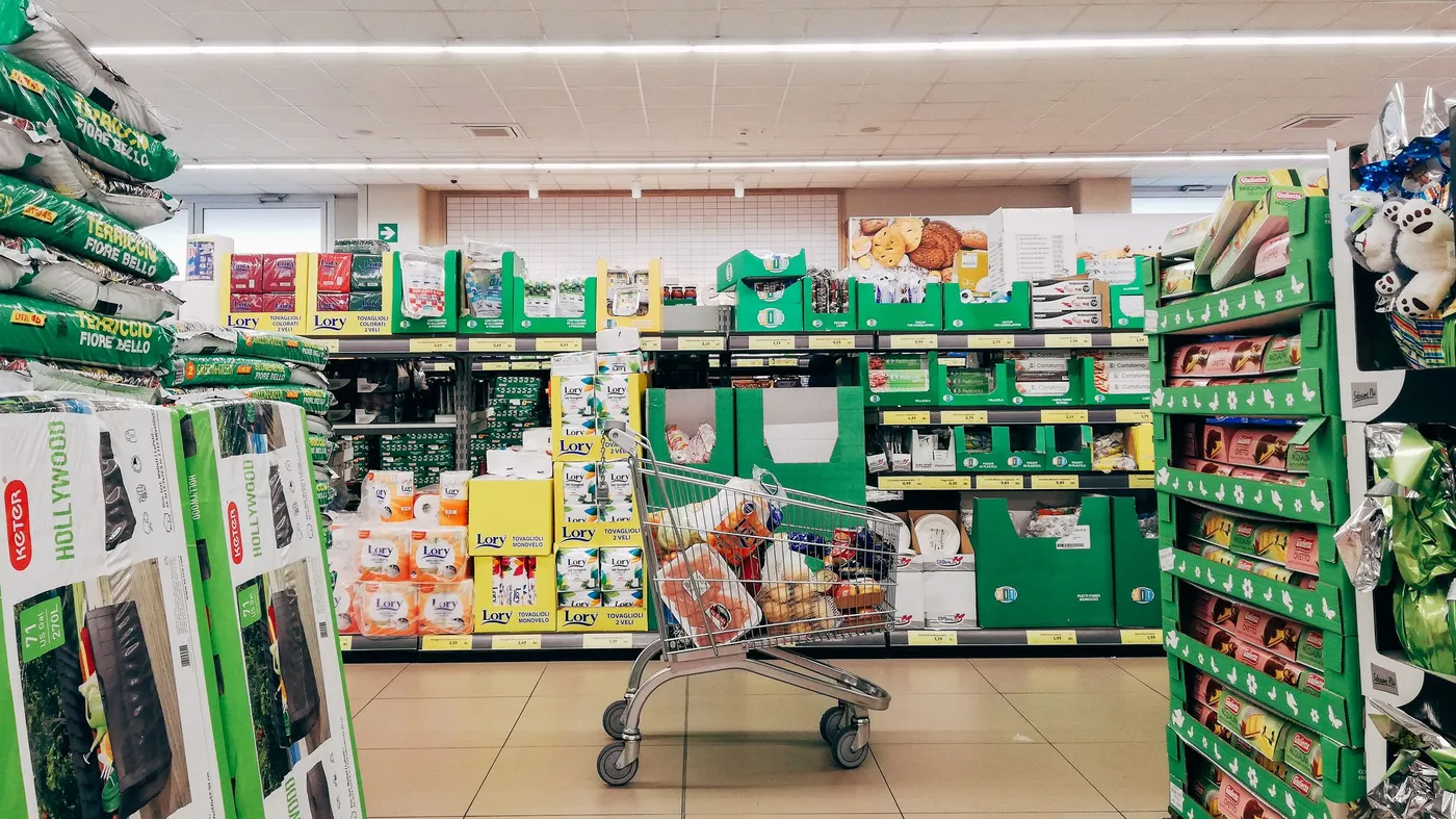 A shopping cart full of groceries stands in the middle of a supermarket aisle lined with green shelves and stacked paper towels.