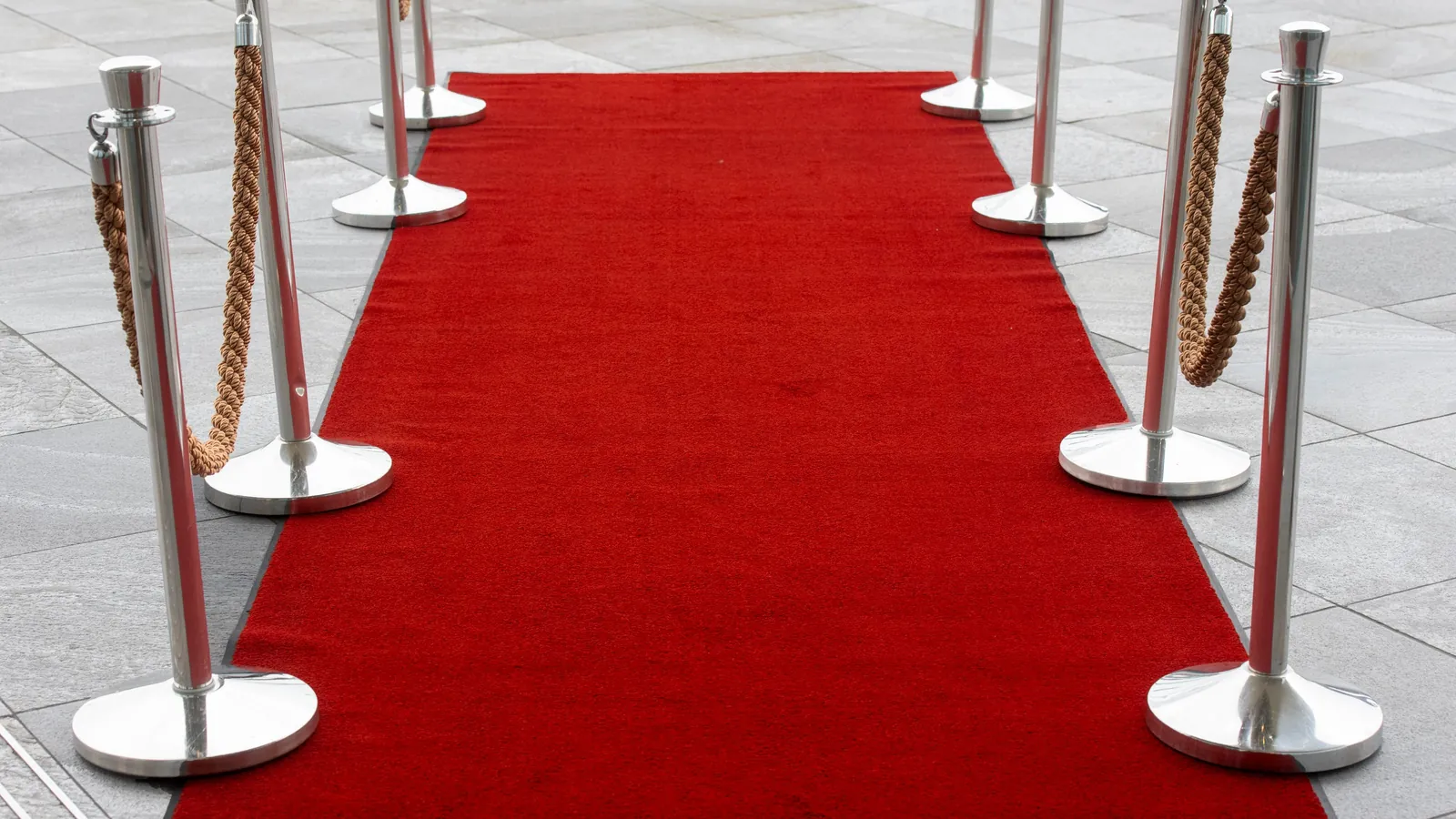 A red carpet running between steel posts and rope barriers on a grey tiled plaza, an entrance open only to people on the list.