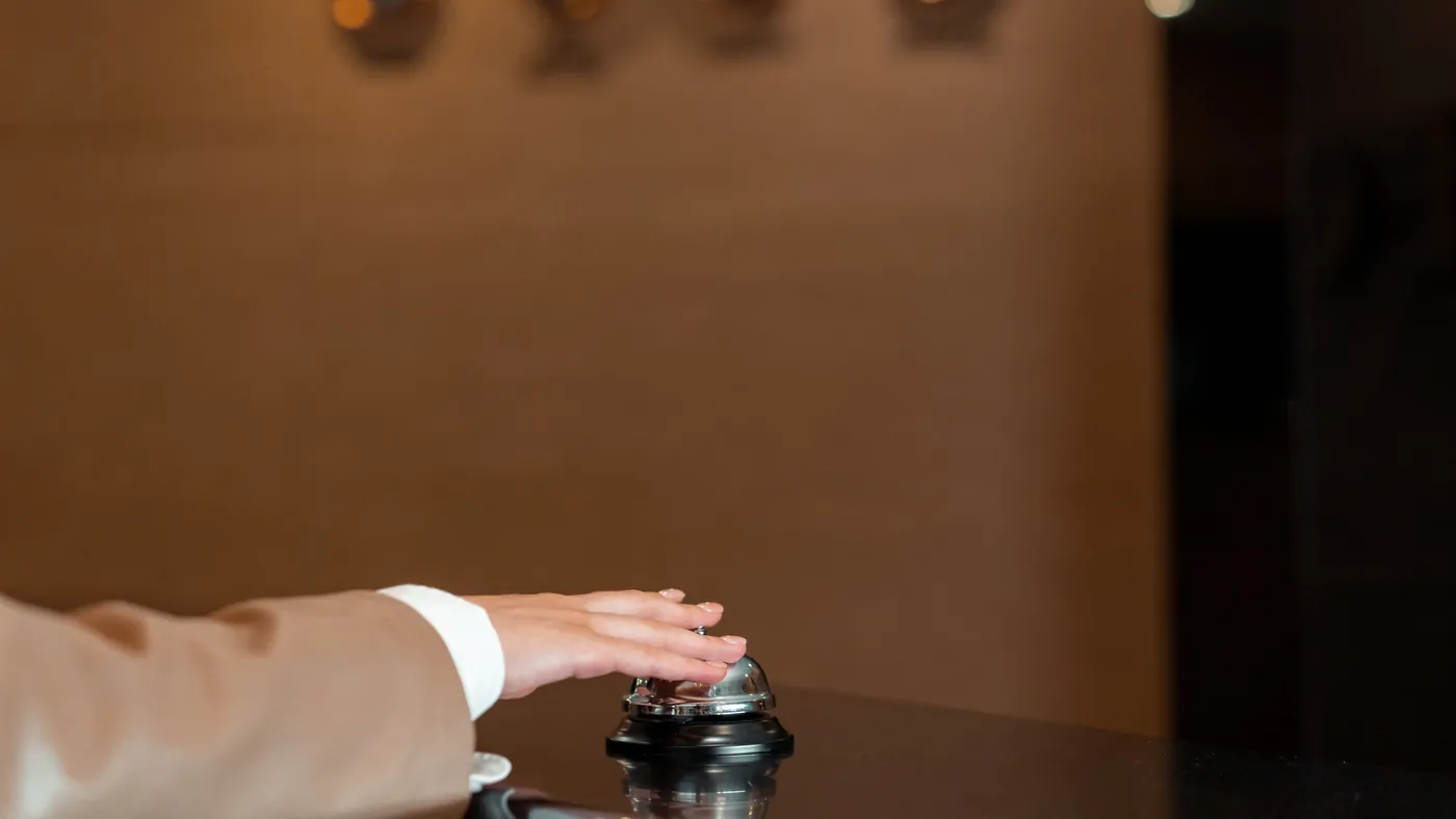 A hand resting on a metal service bell at a dark hotel reception counter, a front desk anyone can walk up to and ring.
