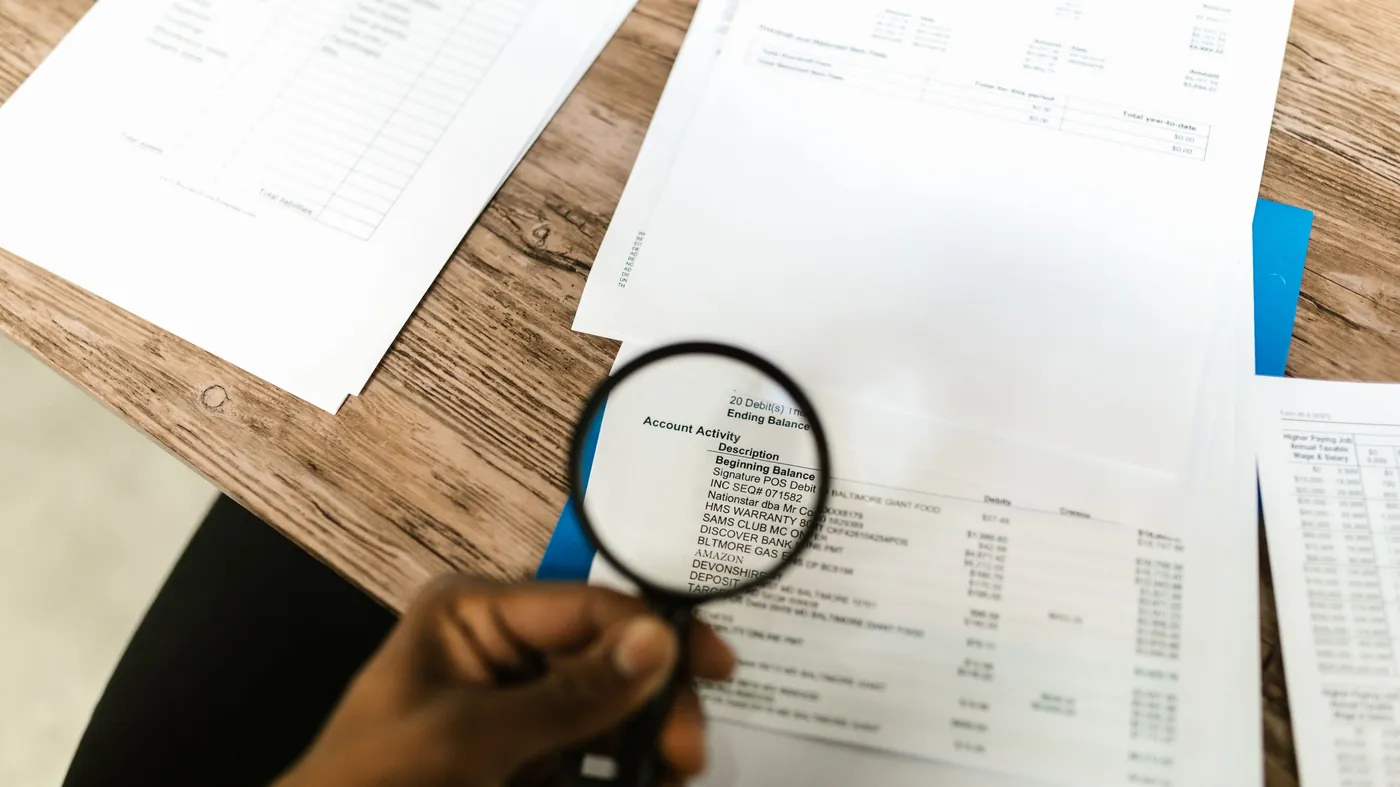 A hand holding a magnifying glass over printed account statements spread across a wooden desk, reading the detail line by line.