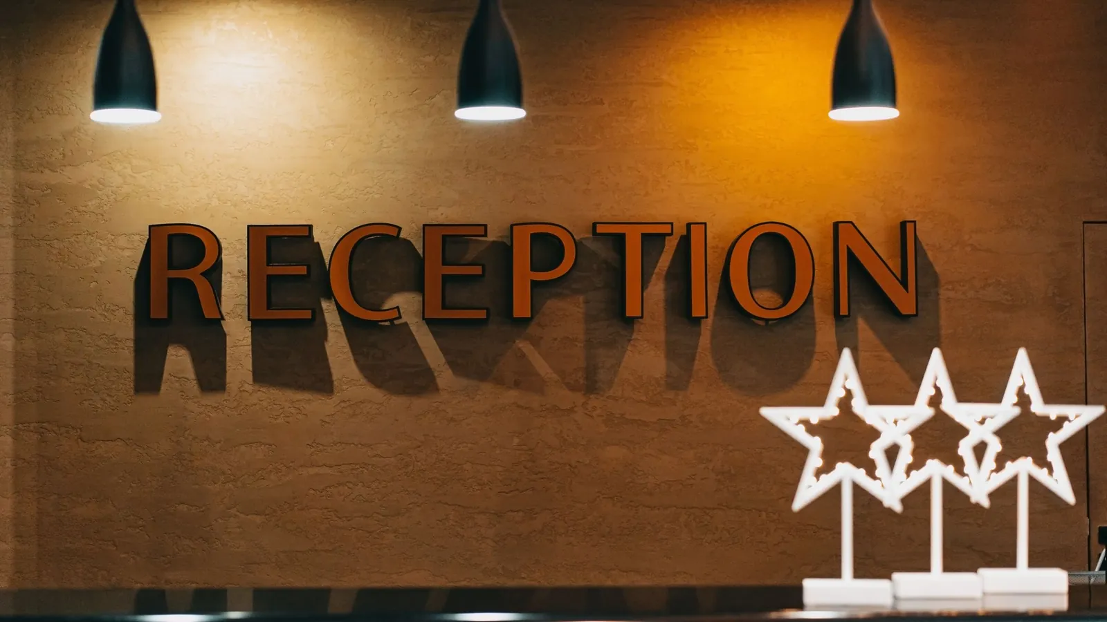 A hotel reception wall with the word RECEPTION in raised letters under three pendant lamps, with lit star decorations on the desk.