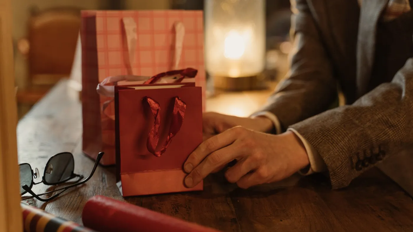 A man in a tweed jacket rests his hand beside red gift bags on a wooden table, with reading glasses and a lamp glowing in the background.