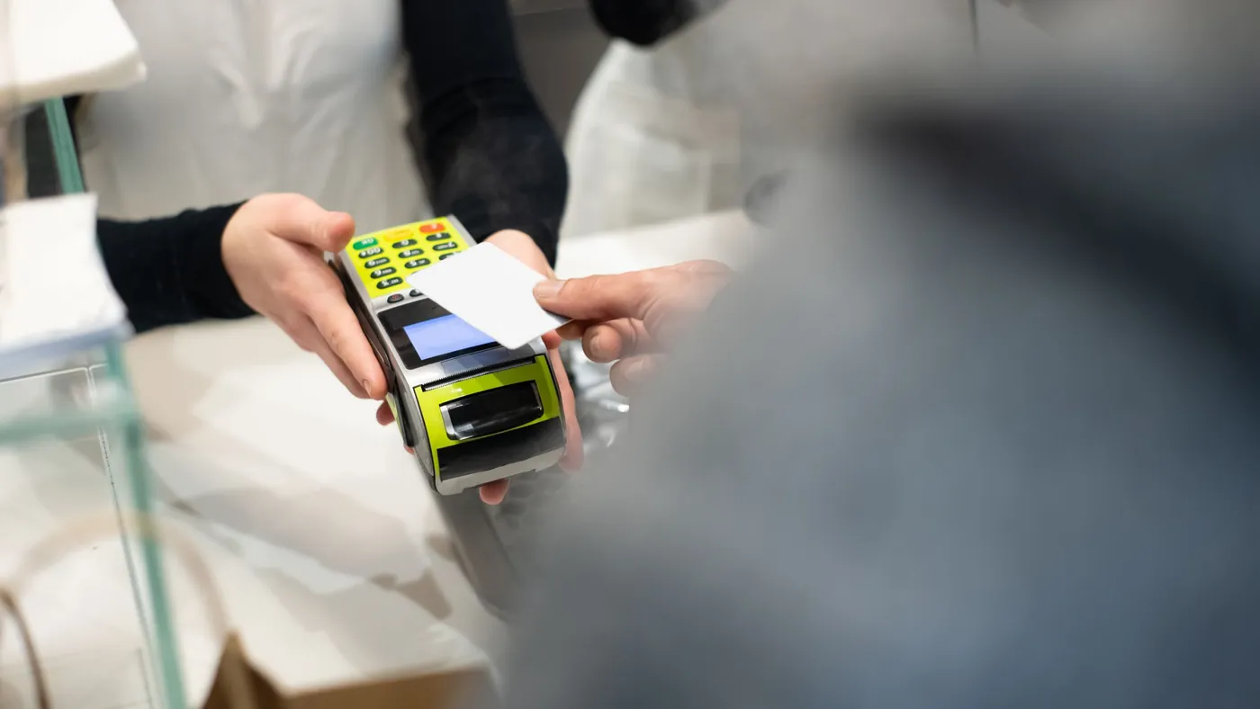 A shop worker holding out a card payment terminal to a customer across a counter, standing in for the authorization step that decides whether an agent-driven purchase completes or stalls.
