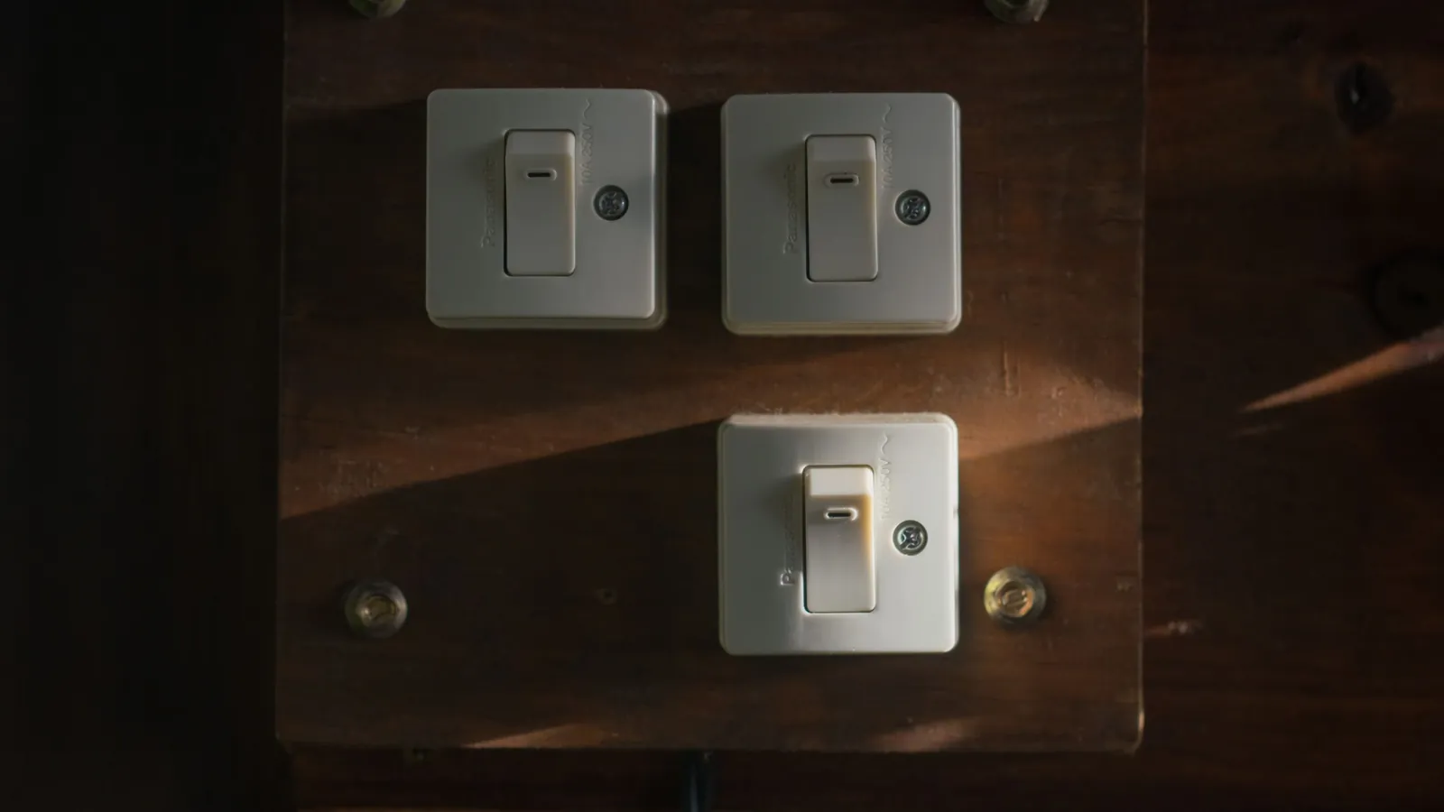 Three old wall switches on a wooden panel in a dim room, lit by a narrow beam of evening light.