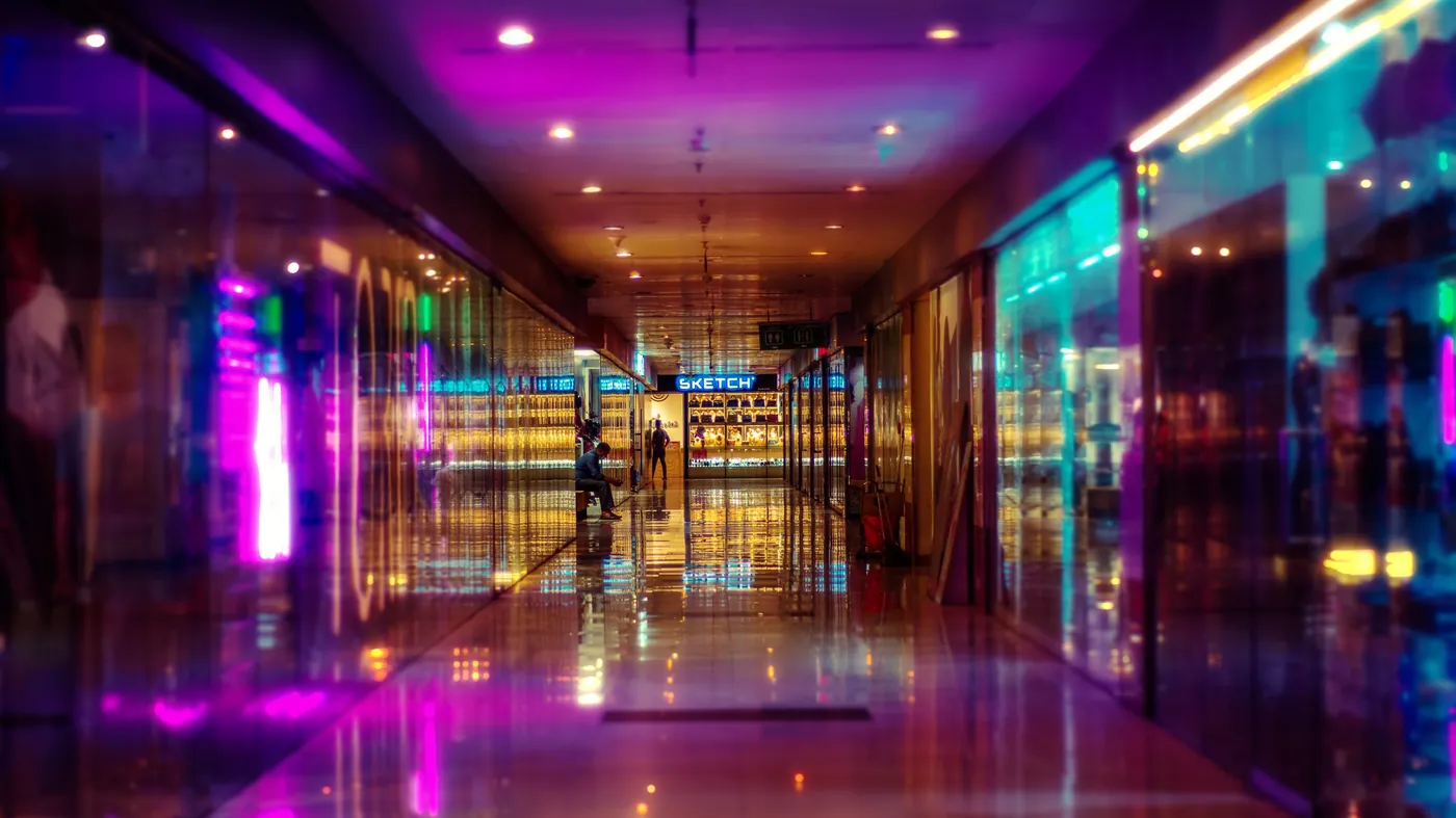 A long shopping center corridor at night lined with brightly lit shop windows reflecting on a polished floor.