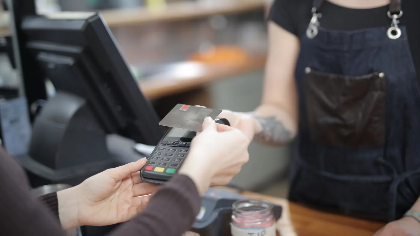 A customer holds a bank card to a payment terminal at a shop counter while the shopkeeper waits.