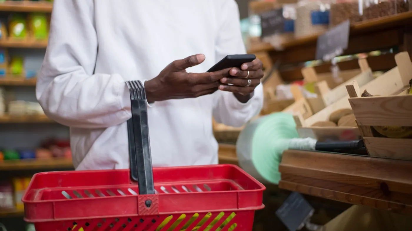 A shopper in a white sweater holds a red shopping basket in one hand and checks a smartphone with the other, beside wooden shelves of goods.