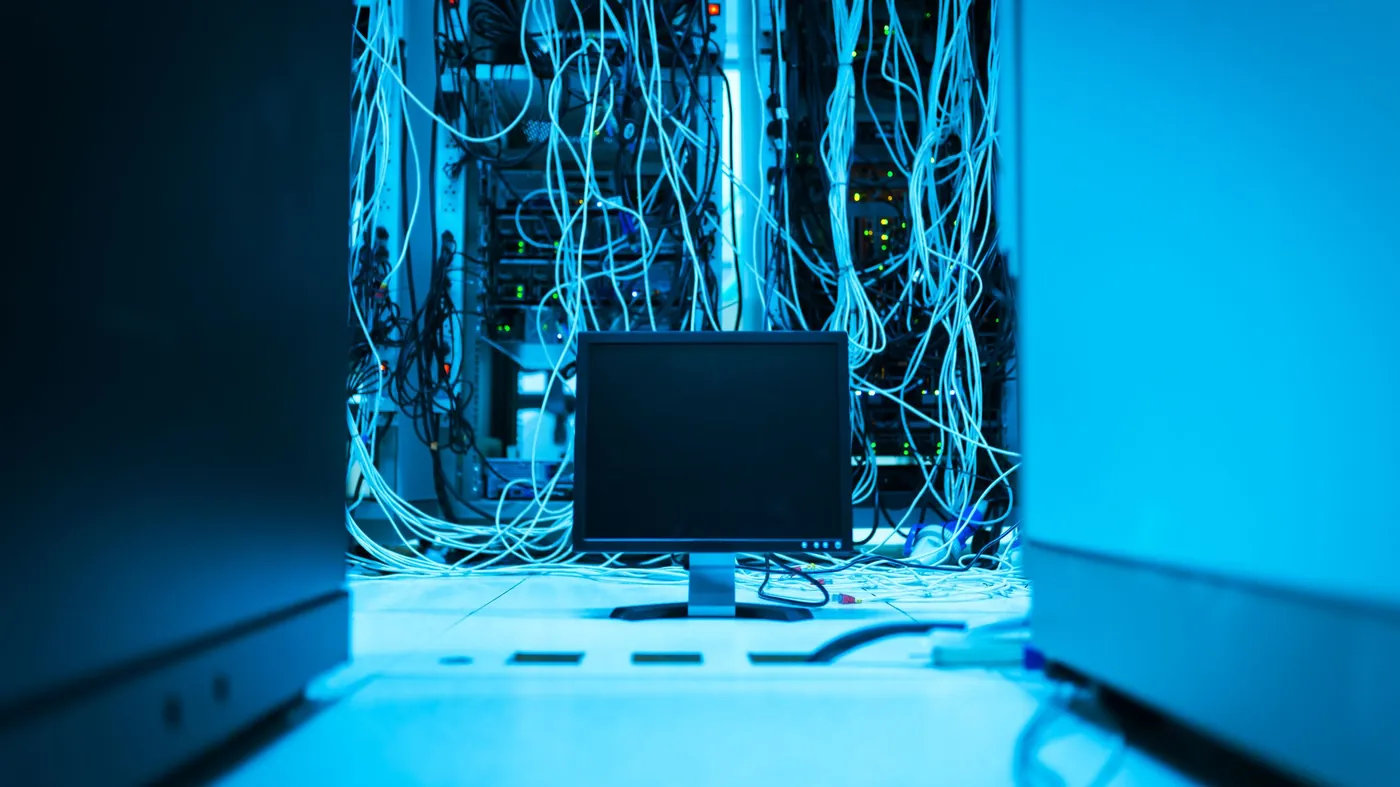 A dark server room lit in blue, with tangled network cables hanging from racks above a single computer monitor.