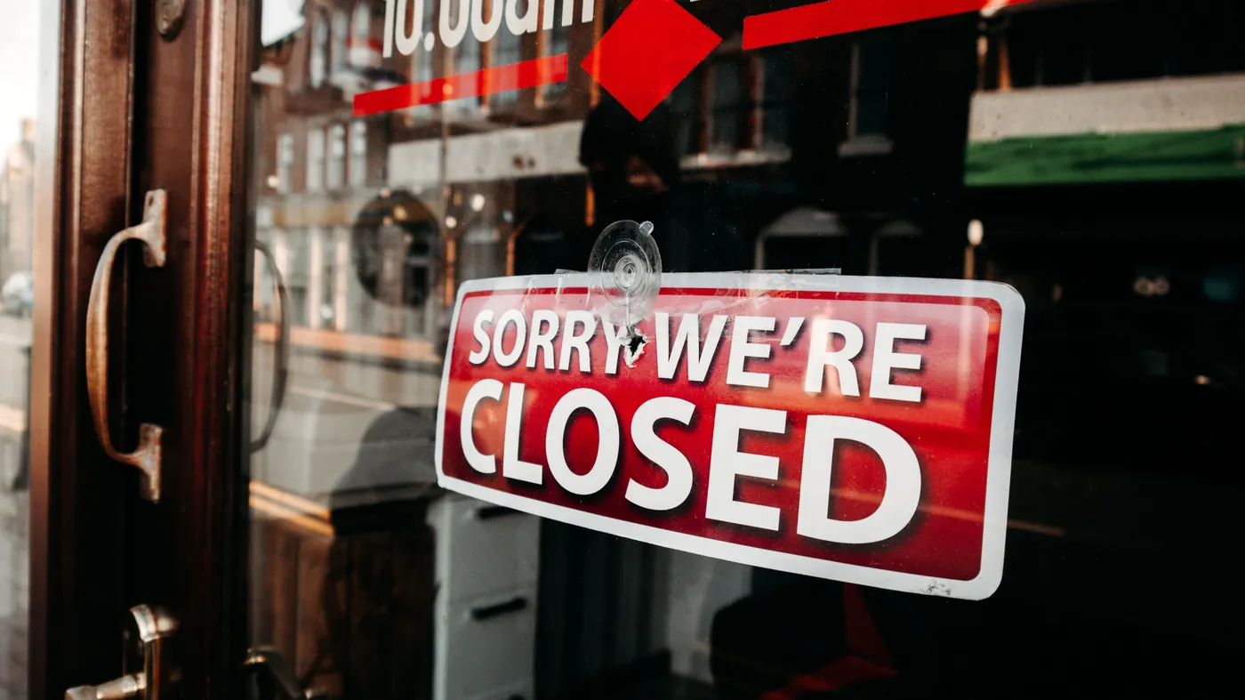 A red Sorry We're Closed sign hangs on the glass door of a shop, with the street reflected behind it.