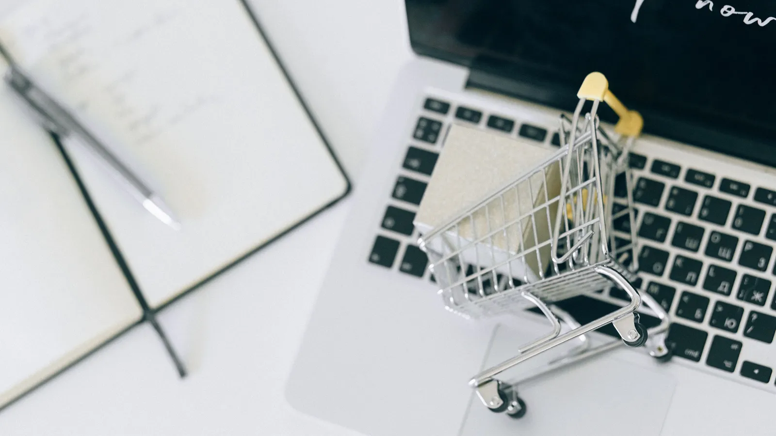 A miniature metal shopping cart holding a small box sits on the keyboard of an open laptop, next to a notebook and pen on a white desk.